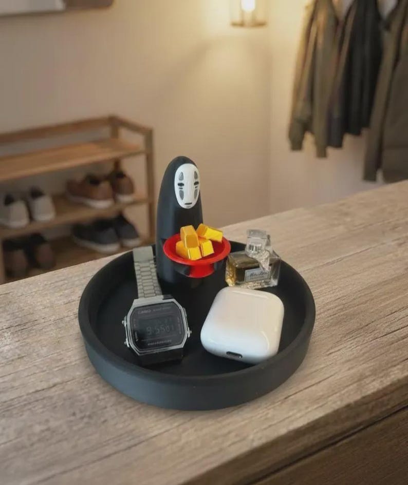 May include: A black round tray holds a digital watch, white wireless earbuds, and small perfume bottles. A black figure with a white face and a red plate with yellow blocks sits on the tray. The tray is on a wooden surface.