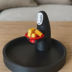 May include: A black decorative tray with a No-Face figure holding a red plate with four yellow blocks. The figure has a white mask and is standing on the tray. The tray is on a wooden surface.
