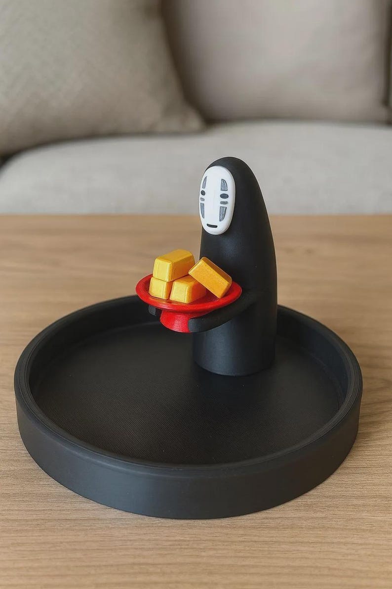 May include: A black decorative tray with a No-Face figure holding a red plate with four yellow blocks. The figure has a white mask and is standing on the tray. The tray is on a wooden surface.