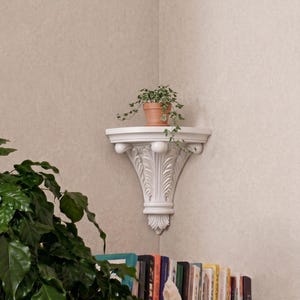 Op de afbeelding: Witte hoekplank met sierlijke details, met een kleine terracotta pot met een groene plant. De plank is aan een neutraal getinte muur bevestigd en biedt een displayruimte voor planten of kleine items.