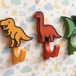Op de afbeelding: Drie wandhaken in de vorm van dinosaurussen. De eerste is een oranje T-Rex, de tweede een rode langnekdinosaurus en de derde een groene pterodactyl. Elke haak heeft een dikke zwarte omlijning en een gebogen basis om voorwerpen aan op te hangen.