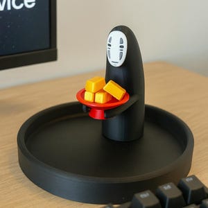 May include: A black figurine of the No-Face spirit from Spirited Away, holding a red tray with four yellow blocks. The figurine is on a black circular tray. A computer keyboard is in the foreground.