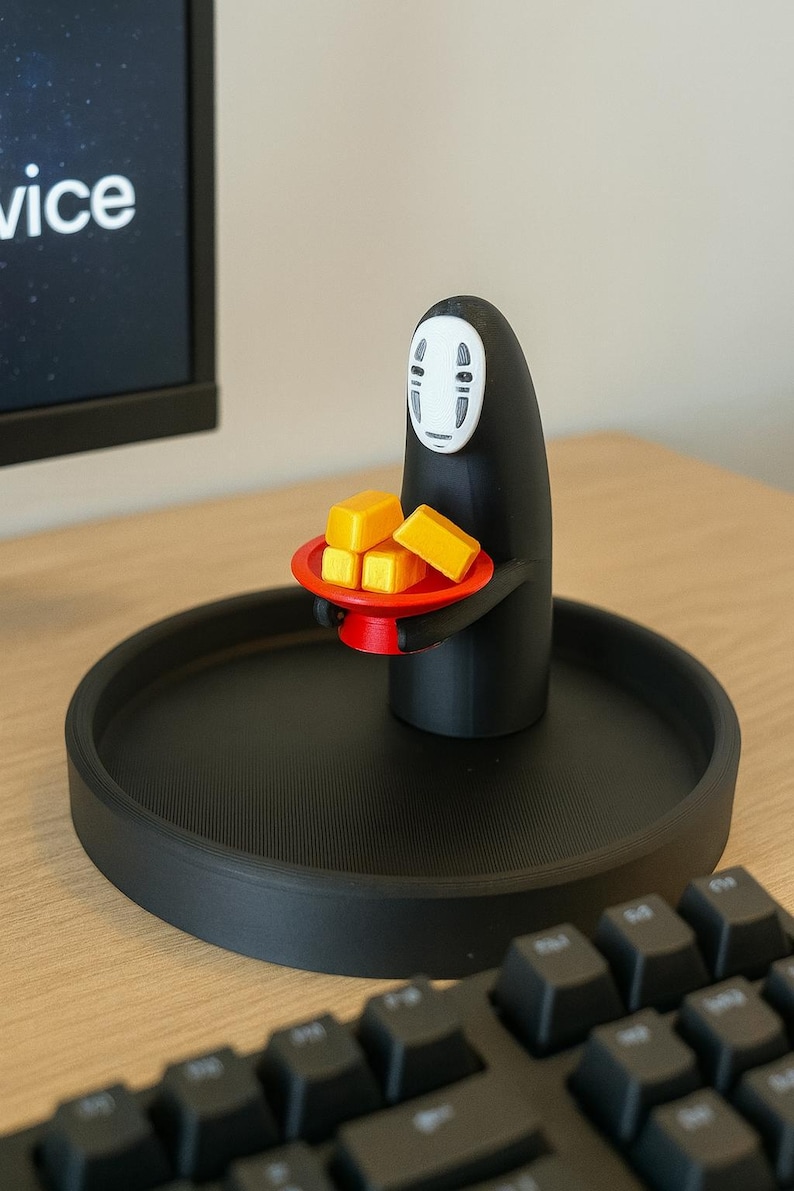 May include: A black figurine of the No-Face spirit from Spirited Away, holding a red tray with four yellow blocks. The figurine is on a black circular tray. A computer keyboard is in the foreground.