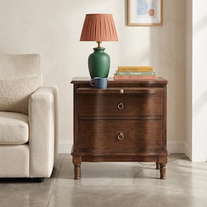 May include: A dark brown wooden nightstand with two drawers, a pull-out tray, and brass ring pulls. On top sits a green ceramic lamp with a pleated terracotta shade, a blue mug, and a stack of three books titled Art & Design.