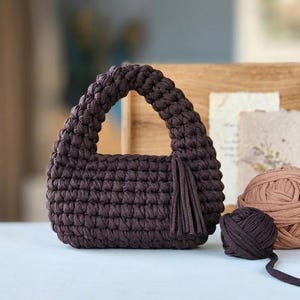 Puede incluir: Un bolso de ganchillo de color marrón oscuro con un asa gruesa integrada y una borla decorativa unida al lateral. El bolso está construido con puntadas de hilo gruesas y uniformes, creando una forma redondeada y texturizada de color sólido.