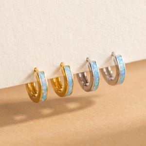 May include: Two pairs of small hoop earrings, one pair in gold-tone metal and one pair in silver-tone metal. Each hoop features a central inlay of iridescent, light blue opal-like material that catches the light with subtle color shifts.