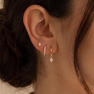 May include: A set of three gold earrings worn in one ear. The top earring is a small gold stud with a four-pointed star shape and a clear stone. The middle earring is a gold hoop with small clear stones. The bottom earring is a gold hoop with a dangling marquise-shaped clear stone charm.