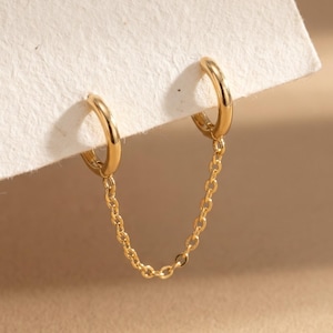 May include: A pair of gold hoop earrings connected by a delicate gold chain. Each small, polished hoop is designed to be worn in two separate piercings, with the fine link chain draping between them to create a continuous, linked jewelry piece.