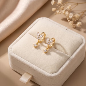 May include: A pair of gold hoop earrings featuring a butterfly-shaped charm adorned with clear, marquise-cut cubic zirconia stones. Each earring includes a small, dangling round cubic zirconia stone suspended from the butterfly design.