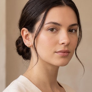 May include: A set of gold-toned earrings consisting of two small hoop earrings and a small stud earring with three tiny clear stones arranged in a row. The jewelry is worn on the earlobe and cartilage, featuring a polished metallic finish.