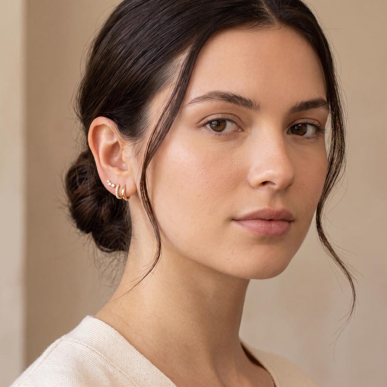 May include: A set of gold-toned earrings consisting of two small hoop earrings and a small stud earring with three tiny clear stones arranged in a row. The jewelry is worn on the earlobe and cartilage, featuring a polished metallic finish.