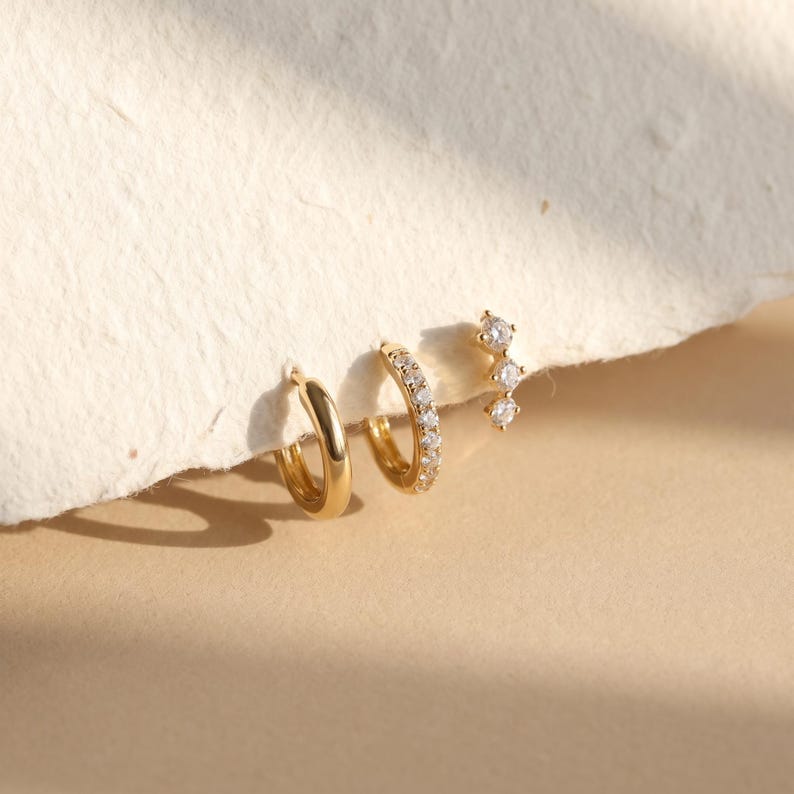 May include: A set of three gold-toned earrings consisting of one plain polished hoop, one hoop encrusted with small clear stones, and one linear stud earring featuring three clear stones arranged in a vertical row, all displayed together.