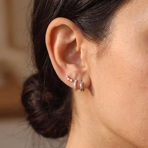 May include: A set of three silver-toned earrings worn in the earlobe. The set includes one small stud earring featuring three tiny clear stones in a row, one small hoop earring encrusted with clear stones, and one plain, smooth silver-toned hoop earring.
