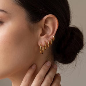 Peut inclure: Un ensemble de cinq créoles dorées graduées portées à une seule oreille. Les boucles d'oreilles présentent un design circulaire lisse et poli, allant d'une grande créole au bas du lobe à quatre créoles progressivement plus petites vers le haut.