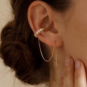 May include: A gold ear cuff featuring a row of small clear stones with a twisted metal border. A delicate gold chain connects the cuff to a thin, straight gold bar earring that hangs vertically from the lower part of the earlobe.