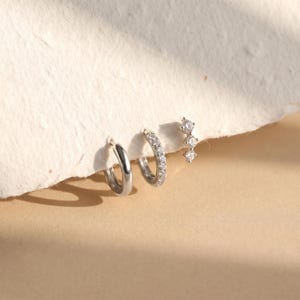 May include: A set of three silver-toned earrings consisting of one plain polished hoop, one hoop encrusted with small clear stones, and one stud earring featuring three vertically aligned clear stones in a graduated, linear arrangement.