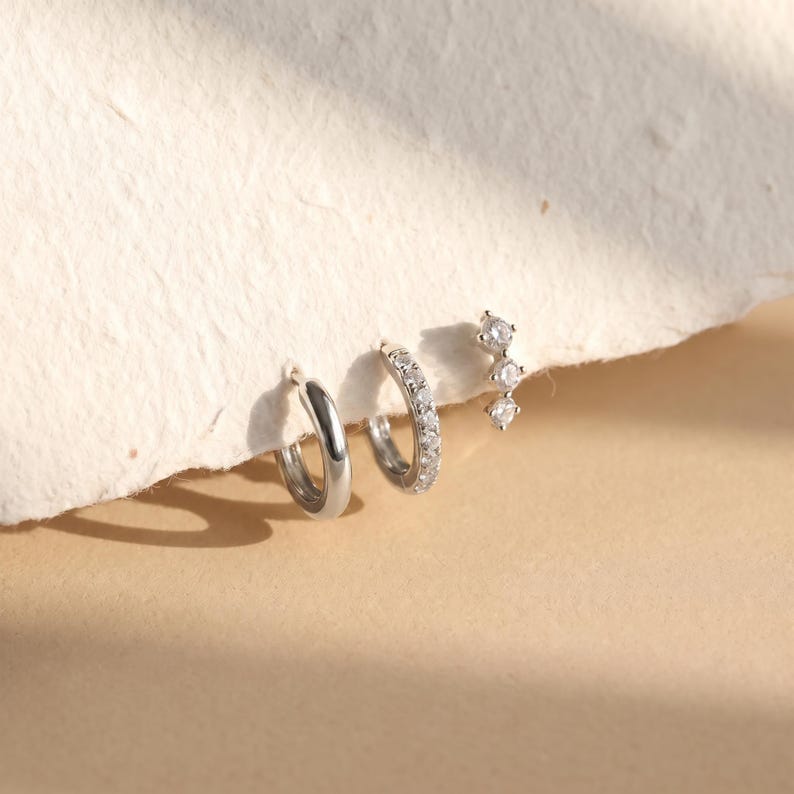 May include: A set of three silver-toned earrings consisting of one plain polished hoop, one hoop encrusted with small clear stones, and one stud earring featuring three vertically aligned clear stones in a graduated, linear arrangement.