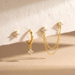May include: A set of three gold-toned earrings featuring a small four-petaled flower stud with clear stones, a small hoop earring with a matching flower charm, and a double-chain earring with two flower studs connected by two delicate gold chains.