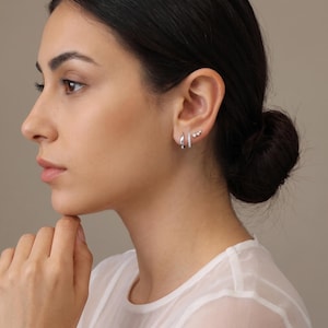 May include: A set of three silver-toned earrings worn in the earlobe and cartilage. The set includes a plain polished hoop, a hoop featuring a row of small clear stones, and a small stud earring with three tiny clear stones arranged in a line.