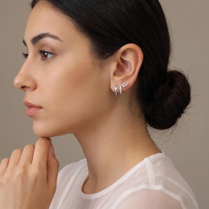 May include: A set of three silver-toned earrings worn in the earlobe and cartilage. The set includes a plain polished hoop, a hoop featuring a row of small clear stones, and a small stud earring with three tiny clear stones arranged in a line.