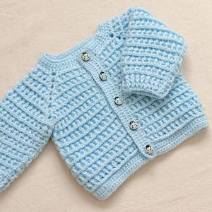 Könnte beinhalten: Hellblauer, handgehäkelter Baby-Cardigan. Der Cardigan hat eine gerippte Struktur, lange Ärmel und wird mit drei Marienkäfer-Knöpfen geschlossen. Das Kleidungsstück ist für Babys konzipiert und aus weichem Garn gefertigt.