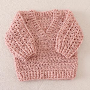 Könnte beinhalten: Ein gehäkelter Pullover in Altrosa mit V-Ausschnitt und langen Ärmeln. Das Kleidungsstück ist mit einem strukturierten Stichmuster an den Ärmeln und einem gerippten Saum gefertigt. Der Hauptteil des Pullovers ist in einem einheitlichen Muster gearbeitet.