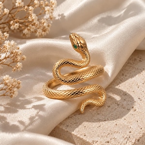 May include: A gold-toned ring shaped like a coiled snake with a textured, scale-like surface and a single small green stone set into the head. The snake body wraps around to form the band, with the head raised and the tail curving downward.