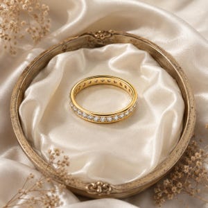 May include: A gold eternity band ring featuring a continuous row of small, clear, round-cut stones set along the entire circumference of the polished metal band, creating a uniform and sparkling appearance around the circular jewelry piece.