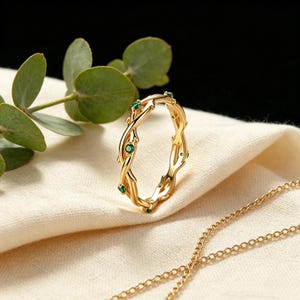 May include: A gold ring featuring a twisted vine design with small, round, green gemstones set at intervals along the band. The ring is positioned upright, showcasing its intricate metalwork and the vibrant color of the embedded stones.