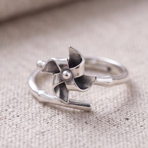 May include: A silver-toned ring featuring a pinwheel-shaped centerpiece with four curved blades and a central rivet. The band of the ring is designed to resemble a segmented bamboo stalk, with an open-ended, adjustable construction.
