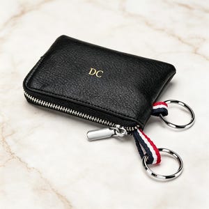 Könnte beinhalten: Schwarzes Leder-Schlüsseletui mit silbernem Reißverschluss und zwei Schlüsselringen. Das Etui hat die Initialen "DC" in goldfarbenen Buchstaben. Eine gestreifte rot-weiß-blaue Stoffschlaufe ist an den Schlüsselringen befestigt.