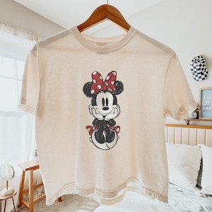 May include: A cream-coloured short-sleeve t-shirt featuring a centred graphic of Minnie Mouse. The character is depicted in a black and white sketch style with a red and white polka-dot bow and matching red and white polka-dot skirt accents.