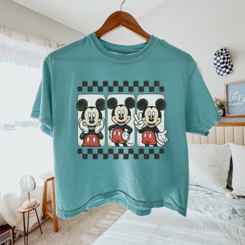May include: A teal short-sleeve t-shirt featuring a graphic print of Mickey Mouse in three different poses within rectangular frames, bordered by black and white checkered patterns above and below the central character illustrations.