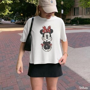 May include: A white short-sleeved t-shirt featuring a graphic print of Minnie Mouse with a red polka-dot bow, paired with a black skirt, a black shoulder bag, and a beige baseball cap. The t-shirt has a relaxed fit and a crew neckline.