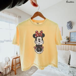May include: A butter yellow short-sleeve t-shirt featuring a distressed graphic print of Minnie Mouse on the front. The character is depicted with her signature red and white polka dot bow, black ears, and hands resting under her chin.