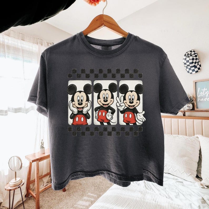 May include: A charcoal gray short-sleeve t-shirt featuring a graphic print of three Mickey Mouse figures in separate panels. The design includes a black and white checkered border above and below the panels, with Mickey in different poses.
