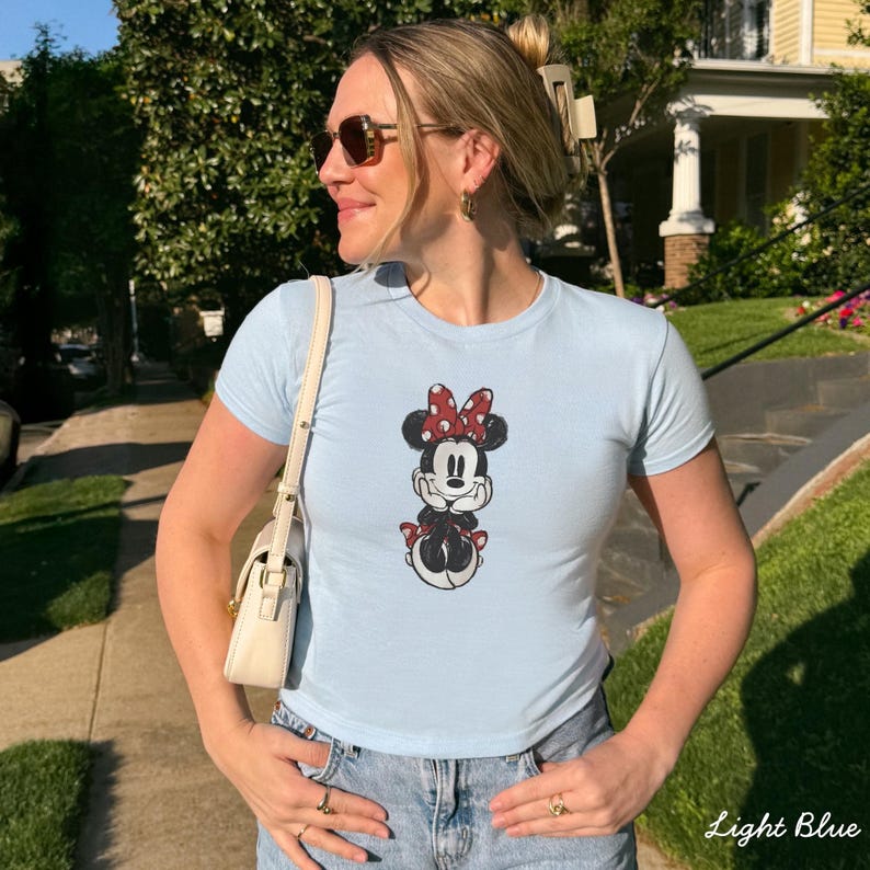 May include: Light blue t-shirt featuring a Minnie Mouse graphic. The cartoon character is depicted with a red bow, black ears, and a red skirt. The shirt is paired with blue jeans and a white purse.