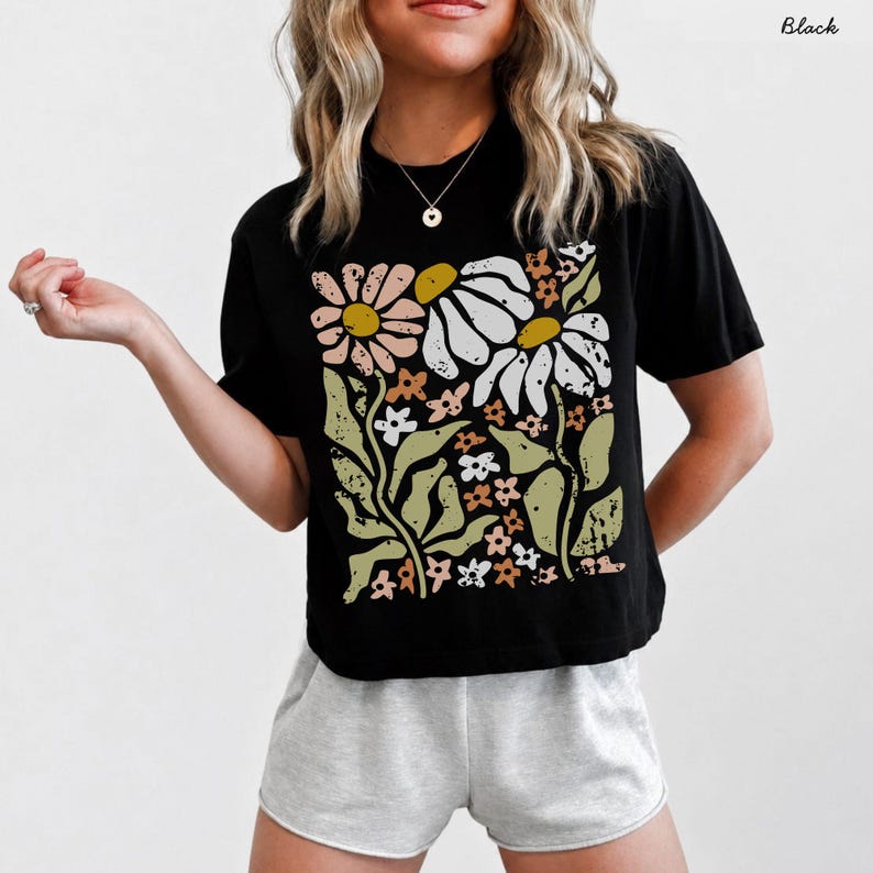May include: A black short-sleeved t-shirt featuring a large, distressed-style graphic print on the front. The design displays a collection of stylized flowers in shades of muted pink, white, mustard yellow, and sage green on a black background.