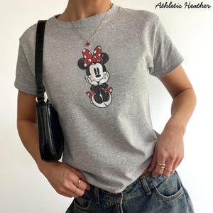May include: Athletic Heather gray t-shirt featuring a Minnie Mouse graphic with a red bow and accents. The shirt is a crew neck style. The text "Athletic Heather" is printed on the top right of the image. The shirt is paired with blue jeans.