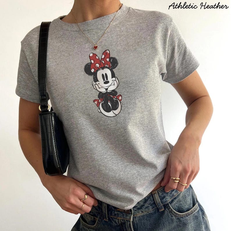 May include: Athletic Heather gray t-shirt featuring a Minnie Mouse graphic with a red bow and accents. The shirt is a crew neck style. The text "Athletic Heather" is printed on the top right of the image. The shirt is paired with blue jeans.