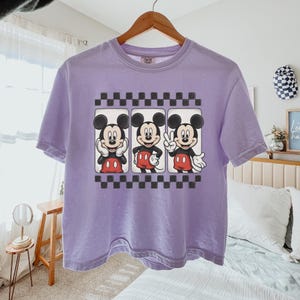 May include: A light purple short-sleeve t-shirt featuring a graphic print of Mickey Mouse in three different poses within rectangular frames, bordered by black and white checkered patterns above and below the central character illustrations.