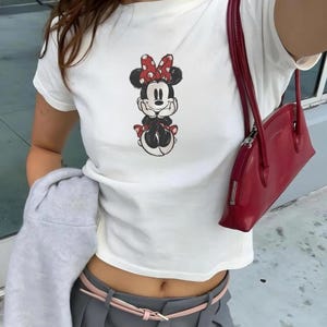 May include: White t-shirt featuring a Minnie Mouse graphic. The cartoon character is depicted in black, red, and white. A red handbag with a zipper closure is visible. The outfit is paired with gray pants and a pink belt.