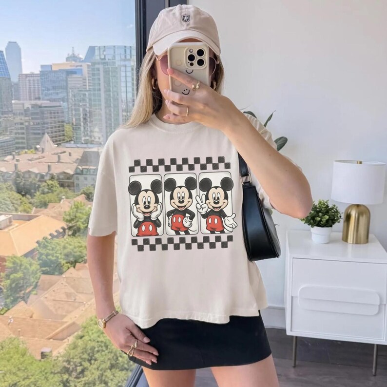 May include: A cream-colored short-sleeve t-shirt featuring a graphic print of Mickey Mouse in three different poses, framed by a black and white checkered border. The shirt is paired with a black skirt, a black shoulder bag, and a beige baseball cap.