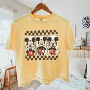 May include: A pale yellow short-sleeve t-shirt featuring a graphic print of Mickey Mouse in three different poses within rectangular frames, bordered by black and white checkered patterns at the top and bottom of the design.