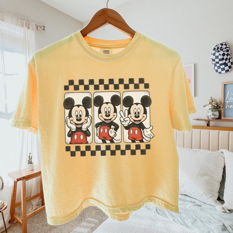May include: A pale yellow short-sleeve t-shirt featuring a graphic print of Mickey Mouse in three different poses within rectangular frames, bordered by black and white checkered patterns at the top and bottom of the design.