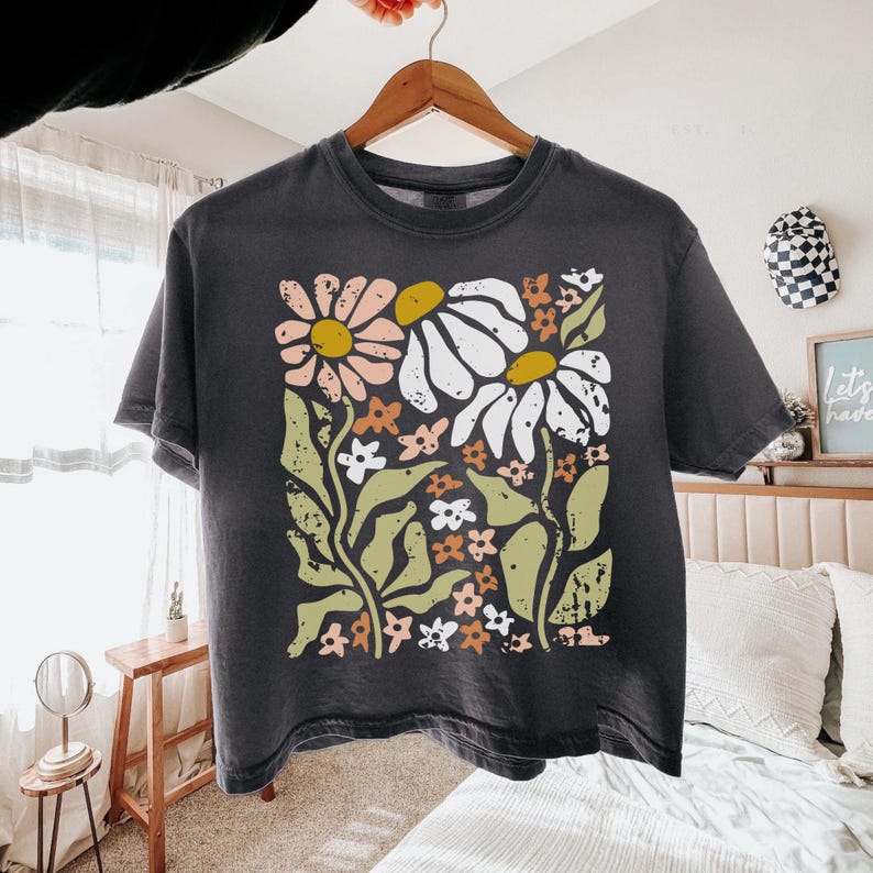 May include: A charcoal gray short-sleeve t-shirt featuring a large, centered graphic print of stylized flowers in shades of muted pink, white, mustard yellow, and sage green. The design has a distressed, textured appearance on the fabric.