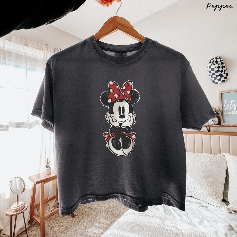 May include: A charcoal grey short-sleeve t-shirt featuring a centred graphic of a cartoon mouse character with a red and white polka dot bow, wearing a red dress and red shoes, with hands resting under the chin in a resting pose.