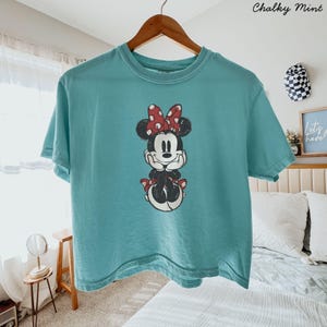 May include: A teal short-sleeved t-shirt featuring a central graphic of Minnie Mouse with her hands under her chin, wearing a red and white polka dot bow. The design has a distressed, vintage-inspired texture on the fabric.