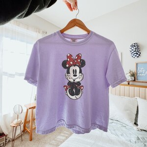 May include: A light purple short-sleeved t-shirt featuring a central graphic of Minnie Mouse with her hands under her chin. The character is wearing a red and white polka dot bow and a matching red and white polka dot skirt. The shirt has a crew neckline.