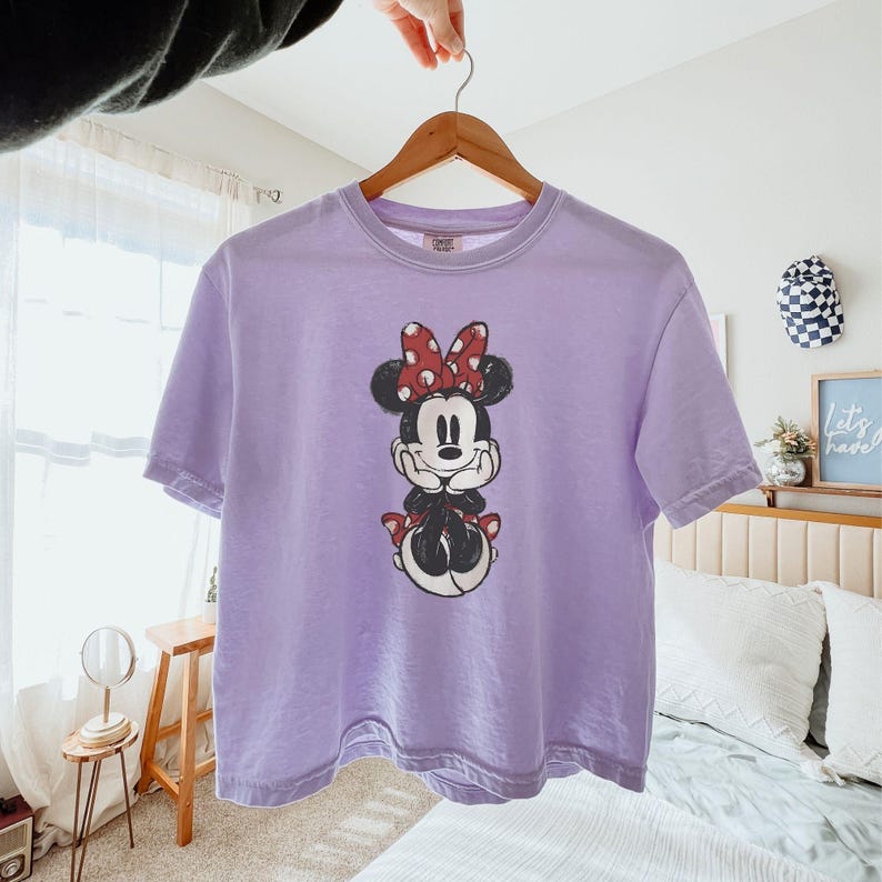 May include: A light purple short-sleeved t-shirt featuring a central graphic of Minnie Mouse with her hands under her chin. The character is wearing a red and white polka dot bow and a matching red and white polka dot skirt. The shirt has a crew neckline.